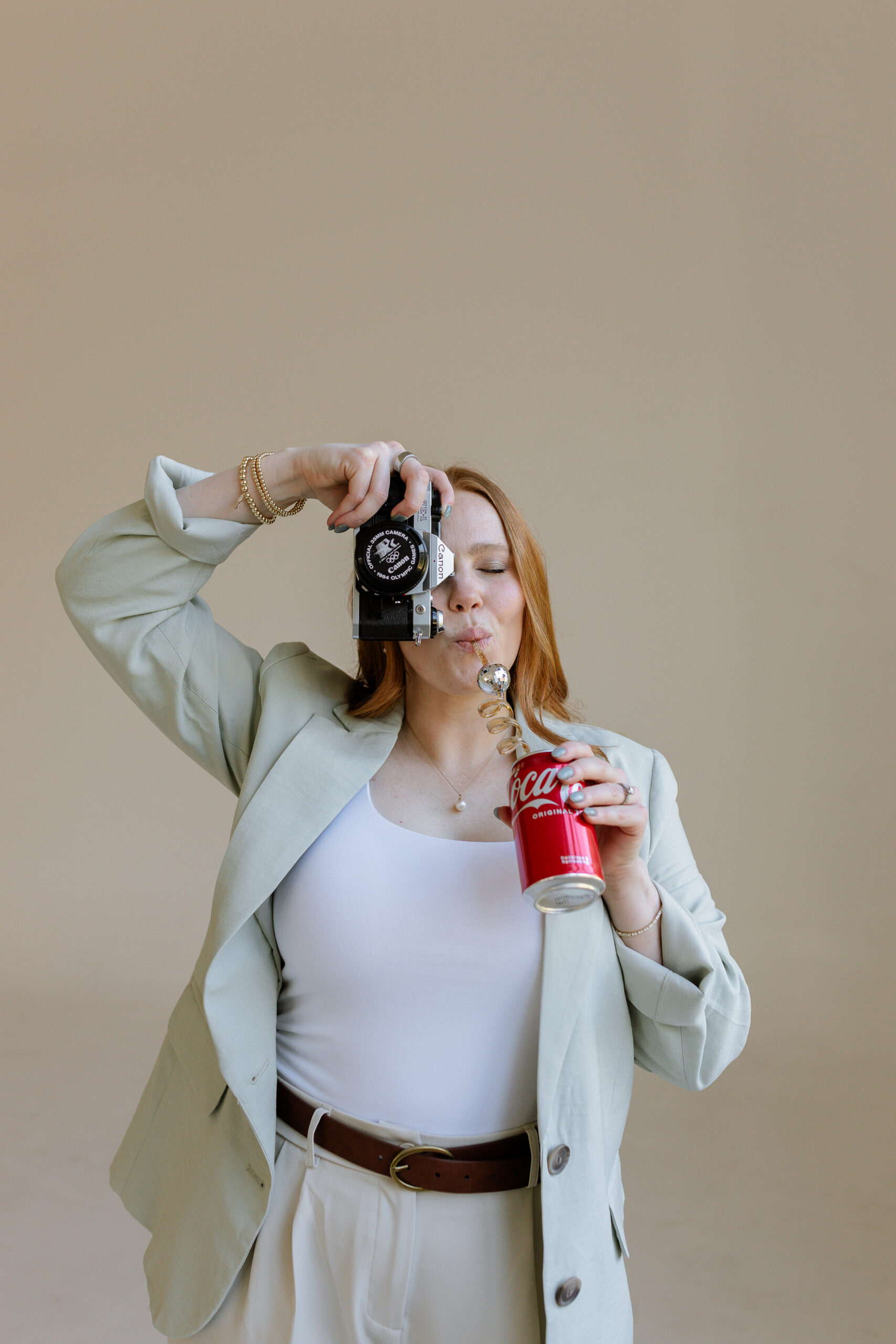 Red-headed woman holding a Canon film camera up to her face and drinking a can of Coke with a twisty straw.